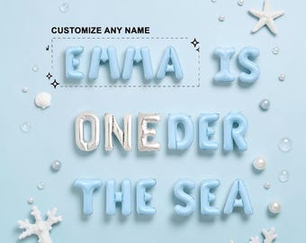 May include: Light blue and silver foil letter balloons spelling out EMMA IS ONEDER THE SEA on a light blue background. The display includes white starfish and coral accents, along with small white pearls scattered across the surface.