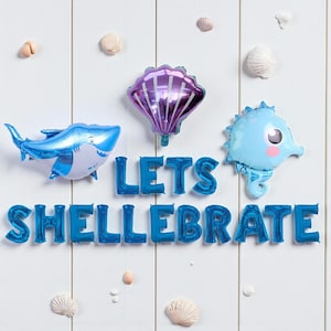 May include: A festive arrangement of blue foil balloons spelling "LETS SHELLEBRATE" with a shark, seashell, and seahorse balloon. White wooden background with seashells.