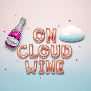 May include: A festive arrangement featuring a pink and silver champagne bottle balloon with the word "Celebrate" and rose gold letter balloons spelling "ON CLOUD WINE". A white cloud-shaped object and confetti complete the celebratory theme.