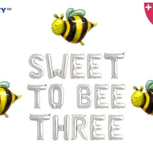 May include: A set of silver foil balloons spelling "SWEET TO BEE THREE" with three yellow and black bee-shaped balloons. The balloons are for a birthday party.