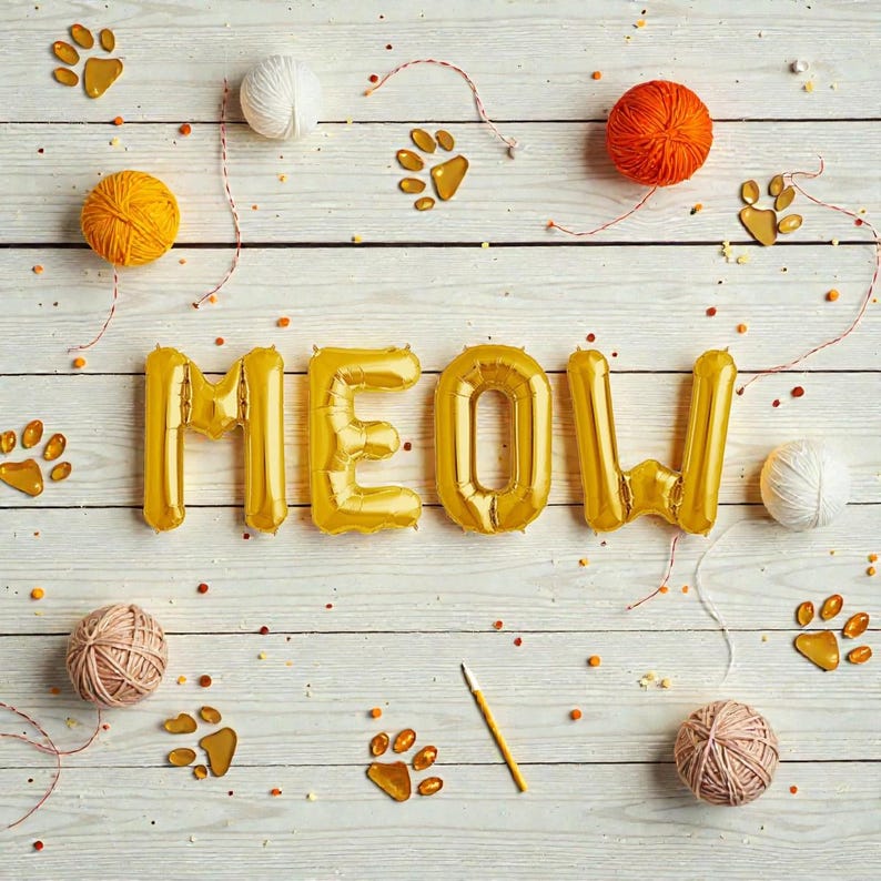 May include: A whimsical arrangement on a white wooden surface featuring gold foil balloon letters spelling "MEOW." Scattered around are orange, yellow, and pink yarn balls, cat paw prints, and a pencil, creating a playful, cat-themed design.