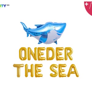 May include: A blue and white shark-shaped foil balloon, with the words "ONEDER THE SEA" in gold foil balloon letters. Includes a balloon straw and string. PartyEight.com logo in the top left corner.