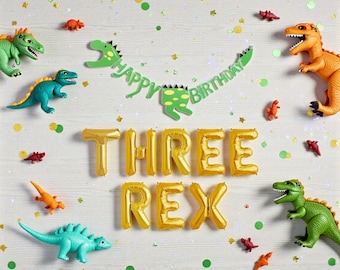 May include: A festive birthday scene with toy dinosaurs and gold foil balloons spelling "THREE REX." A green banner reads "Happy Birthday." Scattered confetti adds to the celebratory atmosphere. Various toy dinosaurs in green, orange, and teal.