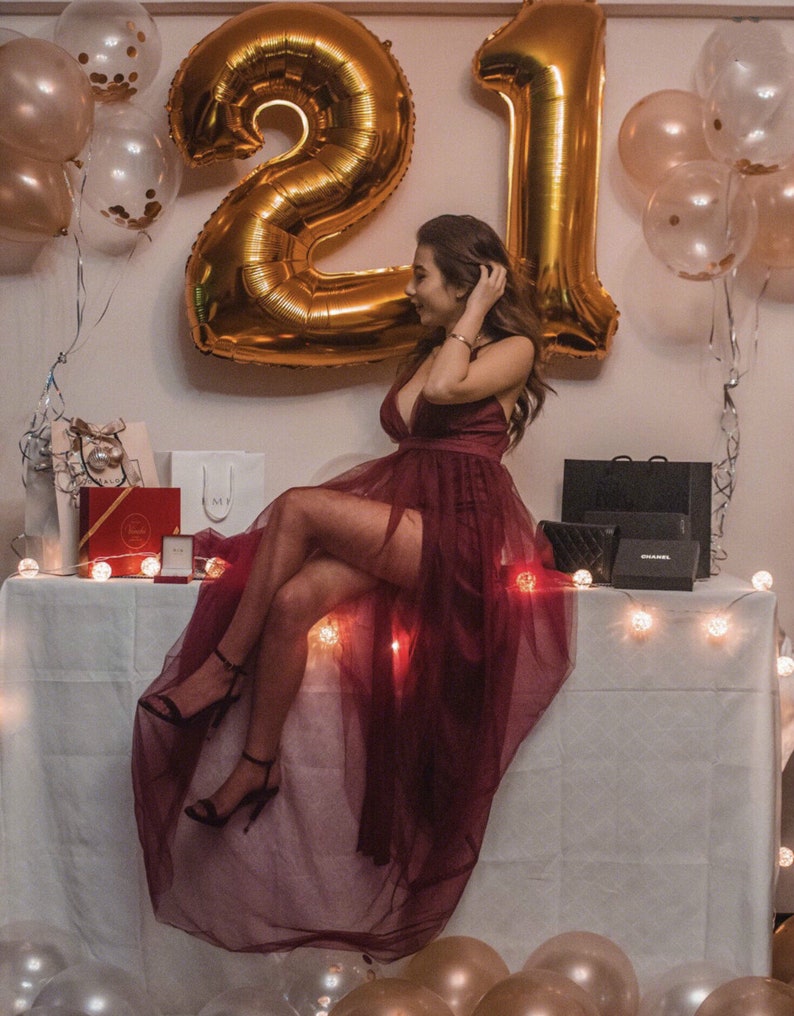 May include: A woman in a burgundy dress with a sheer overlay sits on a table with a white tablecloth.  She is surrounded by gold balloons that spell out "21".