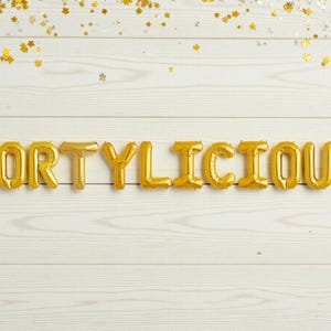 Puede incluir: Letras doradas de globos de papel de aluminio deletrean la palabra "FORTYLICIOUS" sobre un fondo de madera blanca. Confeti dorado en forma de estrella está esparcido en la parte superior. La imagen tiene un tema de celebración, ideal para un cumpleaños.