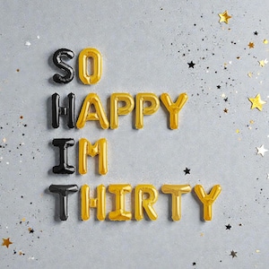 以下が含まれることがあります： 「SO HAPPY I'M THIRTY」と書かれた金と黒の風船文字が特徴的なお祝いの画像。明るい灰色の表面に配置され、金と銀の星型の紙吹雪が散りばめられています。