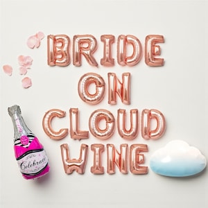 Puede incluir: Globos de papel de aluminio en oro rosa que deletrean "BRIDE ON CLOUD WINE" con un globo de botella de champán y una decoración en forma de nube. Los acentos rosas y blancos completan el tema festivo.