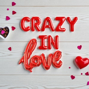 Puede incluir: Escena romántica con letras de globos rojos que deletrean "CRAZY IN LOVE" sobre un fondo de madera blanca. Confeti en forma de corazón y un globo de corazón rojo están dispersos. También hay un cuenco con bombones en forma de corazón.