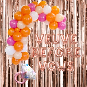May include: A festive party decoration set featuring a balloon arch in shades of orange, pink, white, and peach. Rose gold foil letter balloons spell out "VEUVE BEFORE VOUS" with a diamond ring balloon. The backdrop is a rose gold metallic fringe.