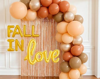 May include: A decorative balloon arch in peach, brown, and gold. The words "FALL IN LOVE" are displayed in gold foil letters. The balloons are arranged in an arch shape, suitable for a party or celebration.