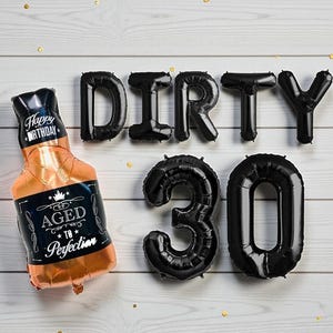 May include: A foil balloon in the shape of a whiskey bottle with the words "Happy Birthday" and "Aged to Perfection." Black balloons spell out "DIRTY 30" on a white wooden background. Gold confetti dots the scene.
