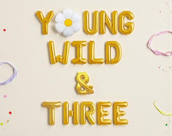 May include: Gold foil balloons spelling "YOUNG WILD & THREE" with a white daisy balloon. The balloons are arranged on a cream background with colorful confetti and ribbons, perfect for a third birthday celebration.