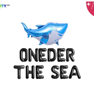 May include: A blue shark-shaped foil balloon with a white belly and a smiling face. Below the balloon, black letter balloons spell out "ONEDER THE SEA". Includes a balloon straw and string.