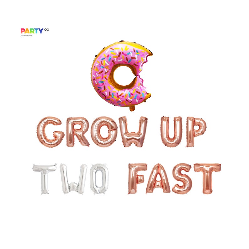 May include: A pink and gold donut-shaped balloon with a bite taken out of it, above rose gold and silver foil letter balloons spelling "GROW UP TWO FAST". Party decorations for a birthday celebration.