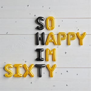 May include: Gold and black foil balloons spelling out "SO HAPPY I'M SIXTY" on a white wooden background. The balloons are arranged in a celebratory display, with silver confetti scattered around.