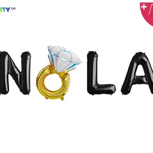 May include: Black foil balloons spelling "NOLA" with a gold ring balloon featuring a large diamond. The balloons are for a party. The image also includes a balloon straw and string.