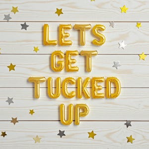 May include: Gold foil balloon letters spell out "LETS GET TUCKED UP" on a white wooden background. Scattered gold and silver star-shaped confetti adds a festive touch to the celebratory scene.
