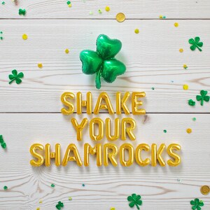 Puede incluir: Imagen festiva del Día de San Patricio con un globo de trébol verde y letras doradas de globo que dicen "SHAKE YOUR SHAMROCKS". Confeti y acentos de trébol dispersos sobre un fondo de vetas de madera blanca.