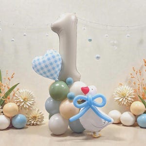 May include: A first birthday decoration with a large number one balloon, a heart-shaped balloon, and a duck balloon. The arrangement includes a cluster of colourful balloons, flowers, and decorative elements, creating a celebratory atmosphere.