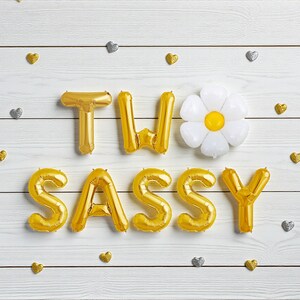 May include: Gold foil balloon letters spelling "TWO SASSY" with a white daisy-shaped balloon and gold and silver heart-shaped confetti on a white wooden background. A festive and celebratory theme.