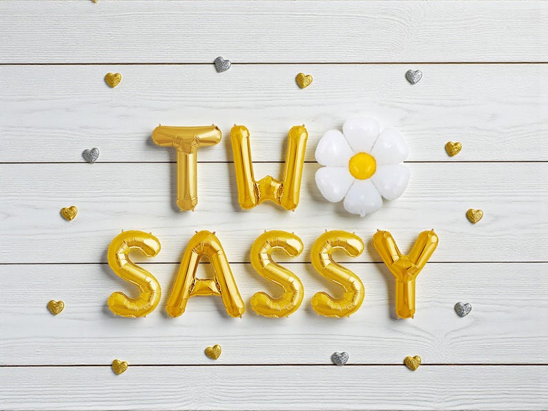May include: Gold foil balloon letters spelling "TWO SASSY" with a white daisy-shaped balloon and gold and silver heart-shaped confetti on a white wooden background. A festive and celebratory theme.