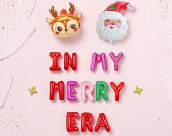 May include: Foil balloons shaped like a reindeer head and Santa Claus face, accompanied by letter-shaped balloons spelling the phrase IN MY MERRY ERA in red, pink, and green, accented by small gold star-shaped balloons on a light pink background.