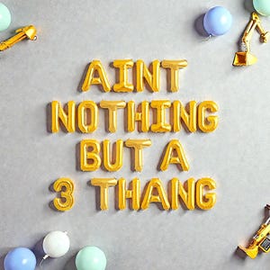 May include: Gold foil balloon letters spell "AINT NOTHING BUT A 3 THANG" on a gray background, surrounded by colorful balloons and toy construction vehicles. A fun and festive party decoration.