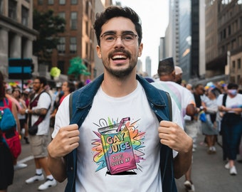 Puede incluir: Una camiseta blanca con un gráfico colorido de una caja de jugo que dice "Gay Juice 100% Queer AF". La persona que lleva la camiseta está sonriendo y de pie en una multitud de personas.