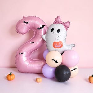 May include: A large pink foil balloon shaped like the number two, decorated with small black bat stickers. Beside it is a white ghost balloon wearing a pink bow and holding an orange pumpkin bucket, accented by a cluster of pink, purple, and black balloons.