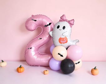 May include: A large pink foil balloon shaped like the number two, decorated with small black bat stickers. Beside it is a white ghost balloon wearing a pink bow and holding an orange pumpkin bucket, accented by a cluster of pink, purple, and black balloons.