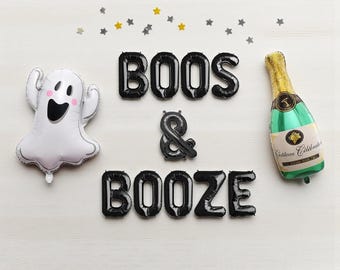 May include: A festive Halloween-themed arrangement featuring a white ghost balloon, black letter balloons spelling "BOOS & BOOZE", and a champagne bottle balloon. Scattered star-shaped confetti adds a celebratory touch.
