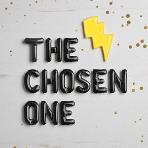 May include: Black balloon letters spell "THE CHOSEN ONE" with a yellow lightning bolt on a white wooden background. Gold confetti dots the surface. The letters are inflated and shiny.
