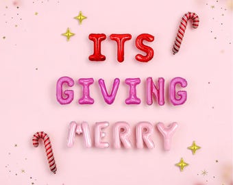 May include: Red and pink foil letter balloons spelling out the phrase ITS GIVING MERRY. The display includes two red and white striped candy cane balloons and four small gold star-shaped balloons arranged on a light pink background.