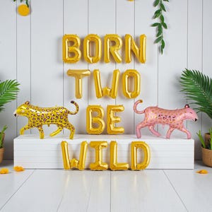K&ouml;nnte beinhalten: Geburtstagsdekoration mit goldenen Folienballons, die "BORN TWO BE WILD" bilden. Zwei Leopardenballons, einer goldfarben und einer rosa, flankieren den Text. Gr&uuml;ne Pflanzen erg&auml;nzen das Party-Thema.