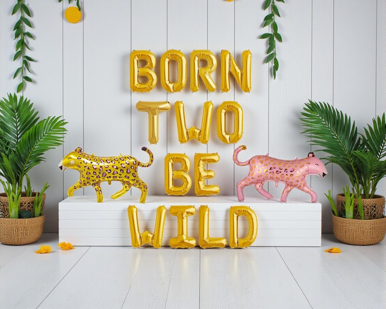 K&ouml;nnte beinhalten: Geburtstagsdekoration mit goldenen Folienballons, die "BORN TWO BE WILD" bilden. Zwei Leopardenballons, einer goldfarben und einer rosa, flankieren den Text. Gr&uuml;ne Pflanzen erg&auml;nzen das Party-Thema.