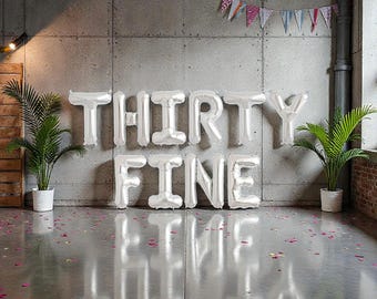 May include: Silver foil balloon letters spell out "THIRTY FINE" against a concrete wall. The floor reflects the text. Two potted plants flank the text, and a banner of triangular flags hangs above. Scattered confetti adds a festive touch.