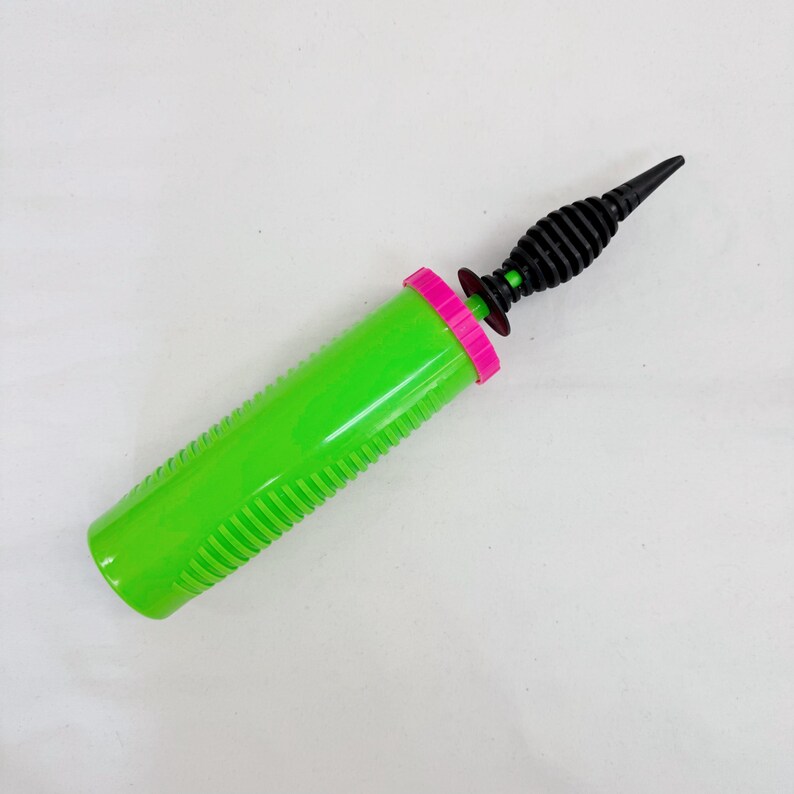 May include: A green and pink manual balloon pump. The pump has a green cylinder with ridged sides, a pink ring, and a black nozzle. The nozzle is black and has a pointed tip. The pump is designed for inflating balloons.