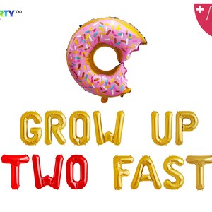 May include: A foil balloon in the shape of a donut, with pink icing and colourful sprinkles, with a bite taken out. Below, gold and red foil balloons spell out "GROW UP TWO FAST". Includes a balloon straw and string.