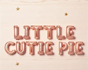 以下が含まれることがあります： 淡いピンク色の木製の背景に、「LITTLE CUTIE PIE」と書かれたローズゴールドの風船文字。 金色の星型の紙吹雪がテキストの周りに散らばっており、お祝いの雰囲気を醸し出しています。