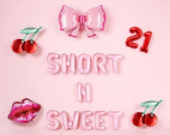 May include: A collection of foil balloons featuring the text SHORT N SWEET in light pink, accompanied by a large pink bow balloon, two red cherry balloons, a red number 21 balloon, and a pink and red lip-shaped balloon on a light pink background.
