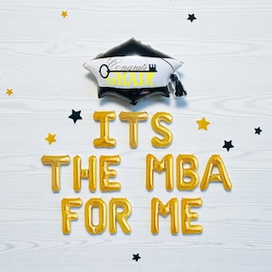 May include: A graduation-themed balloon arrangement with a black and white mortarboard balloon that says "Congrats Grad". Gold foil letter balloons spell out "ITS THE MBA FOR ME" on a white wooden background, with star-shaped confetti.