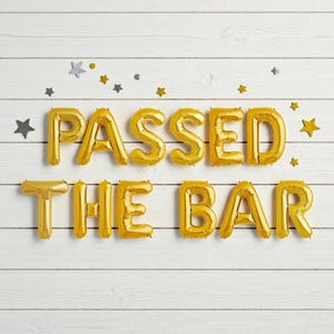 以下が含まれることがあります： 白い木製の背景に、「PASSED THE BAR」と書かれた金色の風船。テキストの周りには、小さな金と銀の星型の装飾が散らばっています。風船は膨らませてあり、2行に配置されています。
