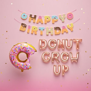 May include: A pink background with a donut-themed birthday banner and balloons. The banner reads "Happy Birthday" with donut-shaped letters. A large, pink donut balloon with sprinkles is on the left, and the phrase "Donut Grow Up" is spelled out in gold balloons.