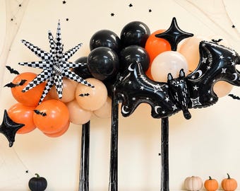 May include: A balloon arch featuring black, orange, and cream-coloured balloons, a large black and white checkered star-shaped balloon, a black bat-shaped balloon with white star and moon prints, and small black bat cutouts attached to the balloons.