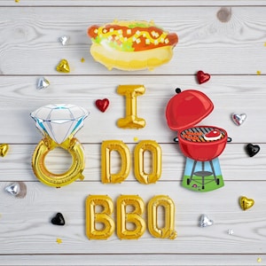 May include: A celebratory display featuring foil balloons and decorations on a light wood surface. The balloons spell "I DO BBQ" and include a diamond ring, hot dog, and a red barbecue grill. Small heart and star-shaped decorations are scattered around.