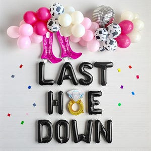 May include: A festive party decoration with a "Last Hoe Down" theme. The arrangement includes pink, white, and cow-print balloons, a disco ball, and black letter balloons. Pink cowboy boot balloons and a diamond ring balloon complete the celebratory scene.