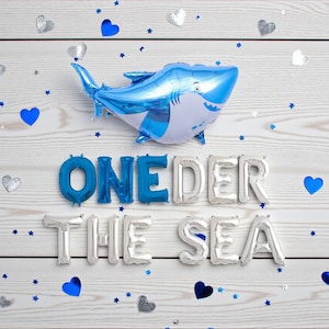May include: A blue and white shark-shaped balloon floats above the words "ONE DER THE SEA" spelled out in silver and blue foil balloons. Scattered around are blue hearts, silver hearts, and blue star-shaped confetti on a white wooden background.