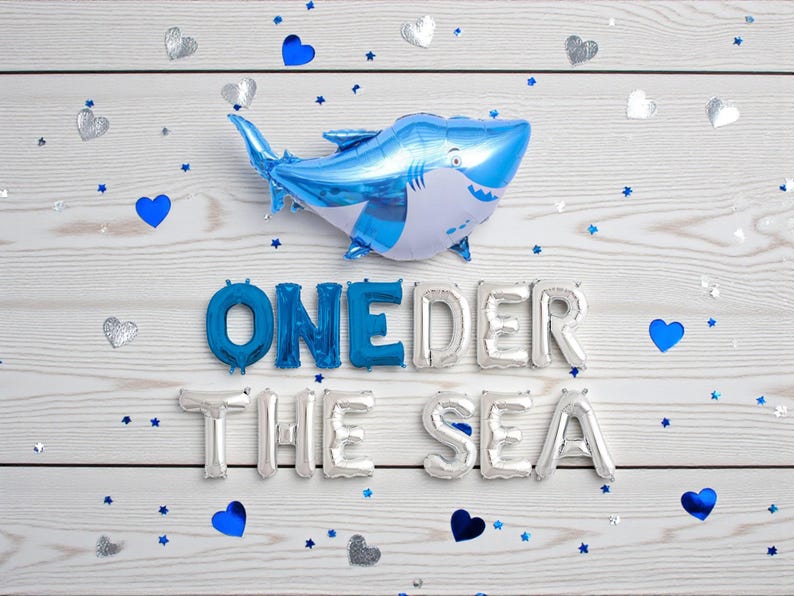 May include: A blue and white shark-shaped balloon floats above the words "ONE DER THE SEA" spelled out in silver and blue foil balloons. Scattered around are blue hearts, silver hearts, and blue star-shaped confetti on a white wooden background.