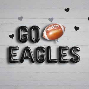 以下が含まれることがあります： 黒い風船の文字で「GO EAGLES」と書かれ、茶色と白のアメリカンフットボールの風船があります。黒とグレーのハート型の装飾が、白い木目調の背景にテキストの周りに散らばっています。