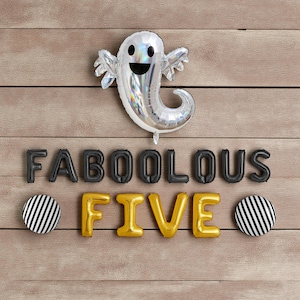 May include: A Halloween display with a silver ghost balloon and the word "FABOOLOUS" in black balloon letters. The word "FIVE" is in gold, with two black and white striped circles. The arrangement is set against a wooden background.